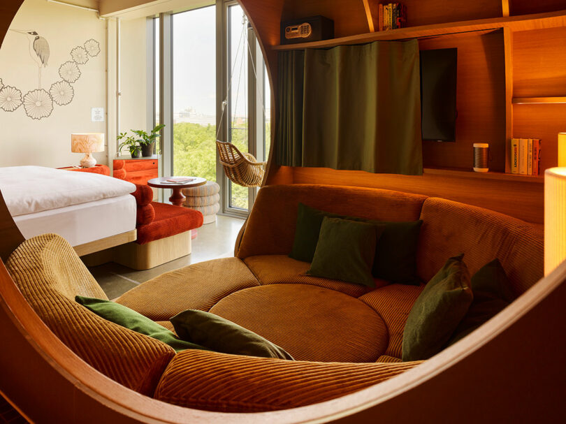 Modern 25hours hotel room with a round sunken seating area, bed, large windows, hanging chair, built-in bookshelf, and warm wood accents.