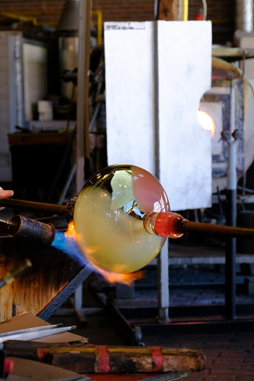 A glassblower shapes a molten glass bubble on a blowpipe in a workshop, with a furnace and tools visible in the background.
