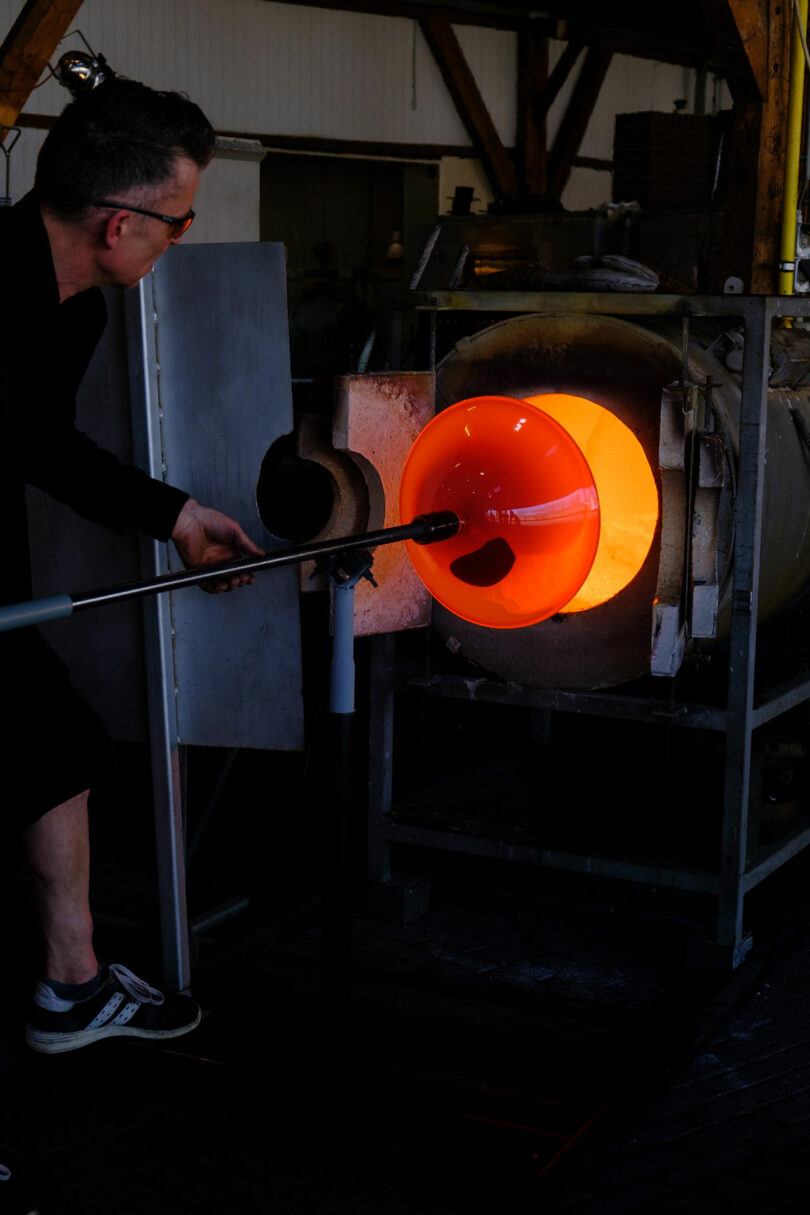 A person shapes molten glass on a blowpipe, rotating it in front of a furnace in a glassblowing workshop.
