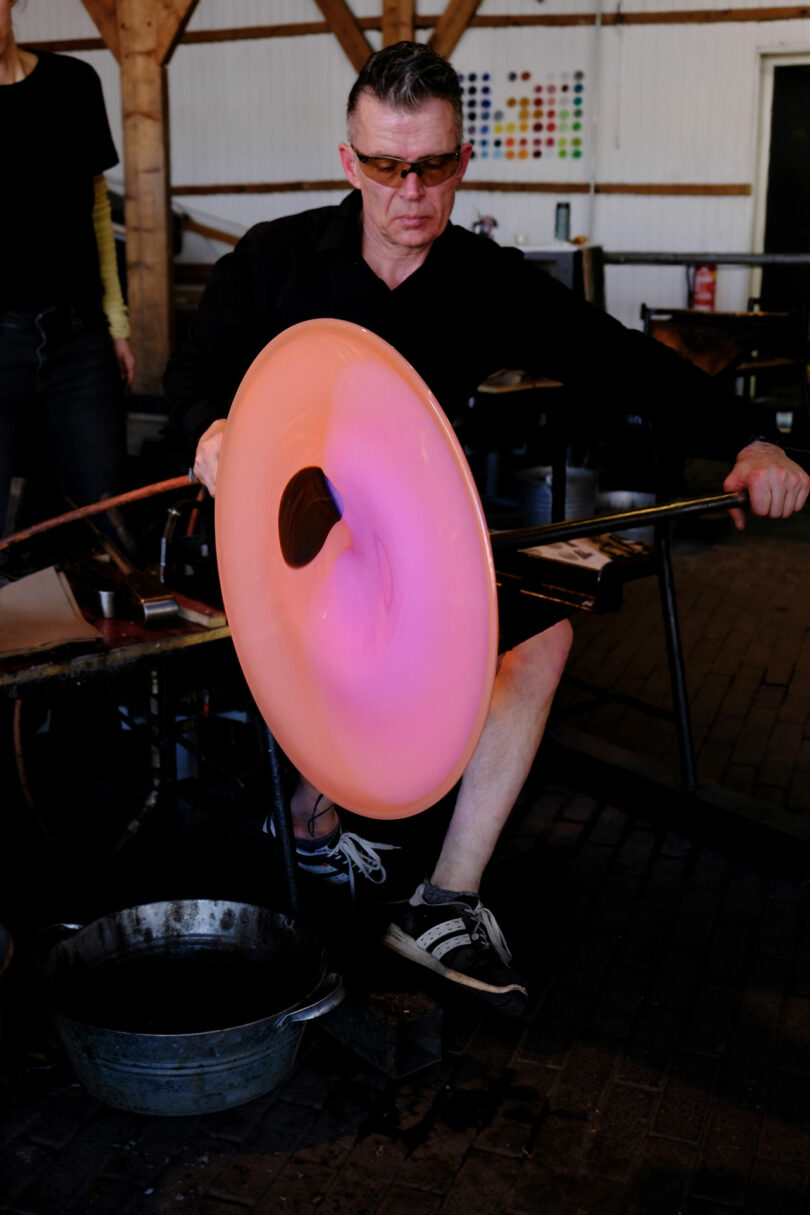 A person in safety glasses shapes a large, glowing pink glass disc with a blowpipe in a workshop setting.