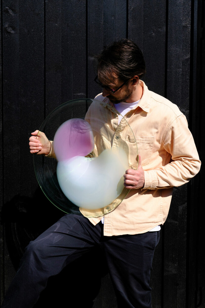 A person in a beige shirt and dark pants holds a transparent circular object with colorful, cloud-like shapes in front of a black wooden wall.
