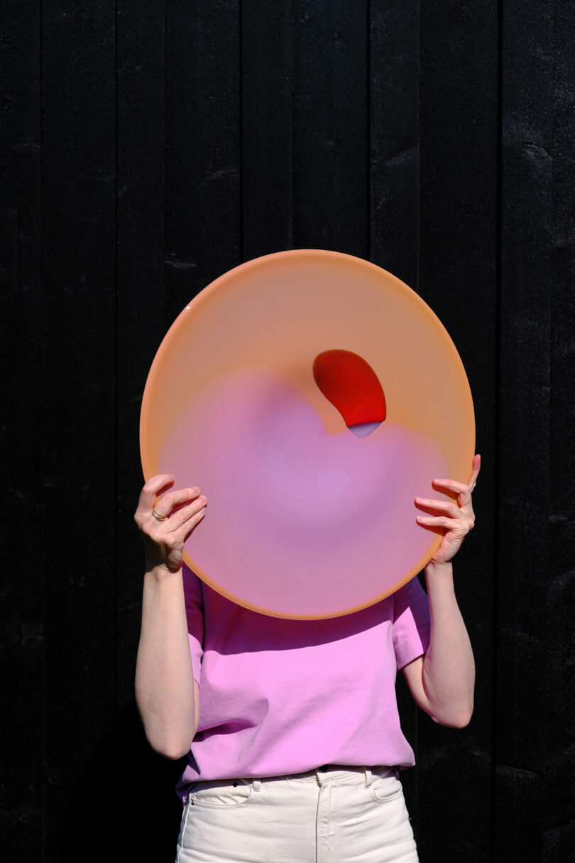 A person in a pink shirt and white pants holds a large, round, peach-and-purple object in front of their face against a black wooden background.