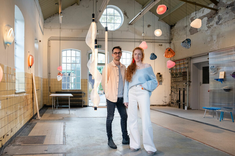 Two people stand in an industrial-style room with high ceilings, exposed pipes, and various modern hanging light fixtures.
