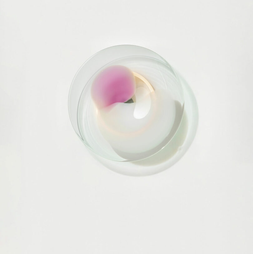 A round, translucent glass object with soft gradients of pink, green, and white, casting a subtle shadow on a white background.