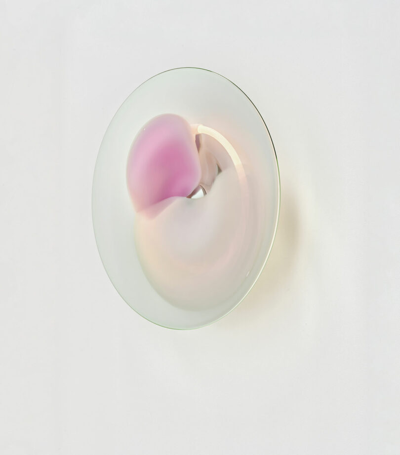 A round, translucent glass object with soft pink and cream hues sits on a white background, casting subtle shadows and reflections.