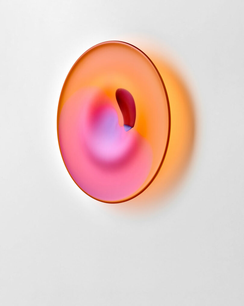 A round, translucent wall sculpture displays gradients of orange, pink, and purple hues with a small, curved, darker element near its center, mounted on a white background.