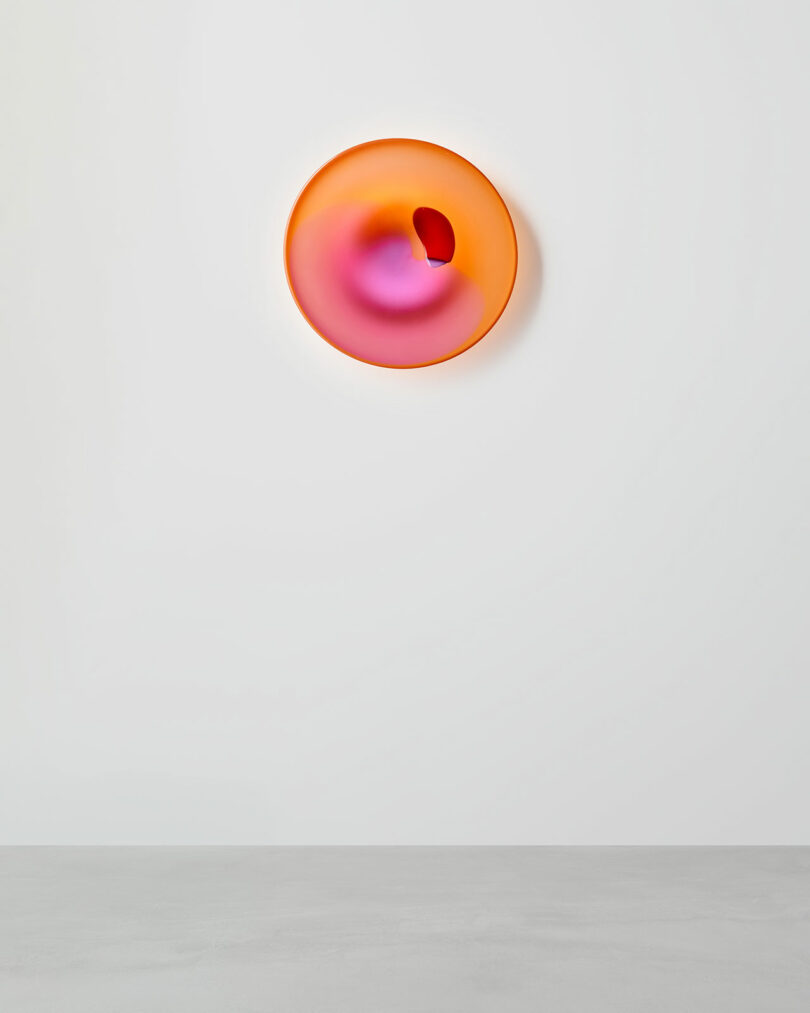 A circular, wall-mounted abstract sculpture with a smooth gradient of orange, pink, and purple tones, featuring a central fold or indentation.