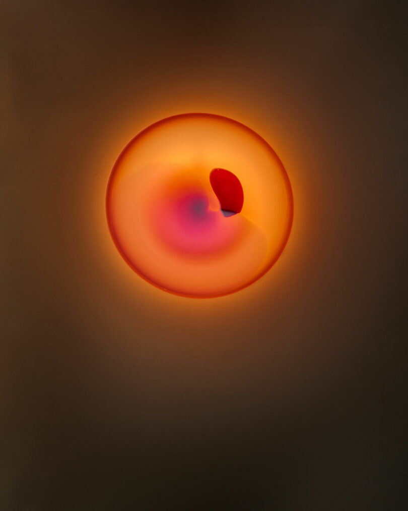 A circular abstract shape with a glowing orange and pink gradient center, surrounded by a dark background and featuring a red crescent form inside the circle.