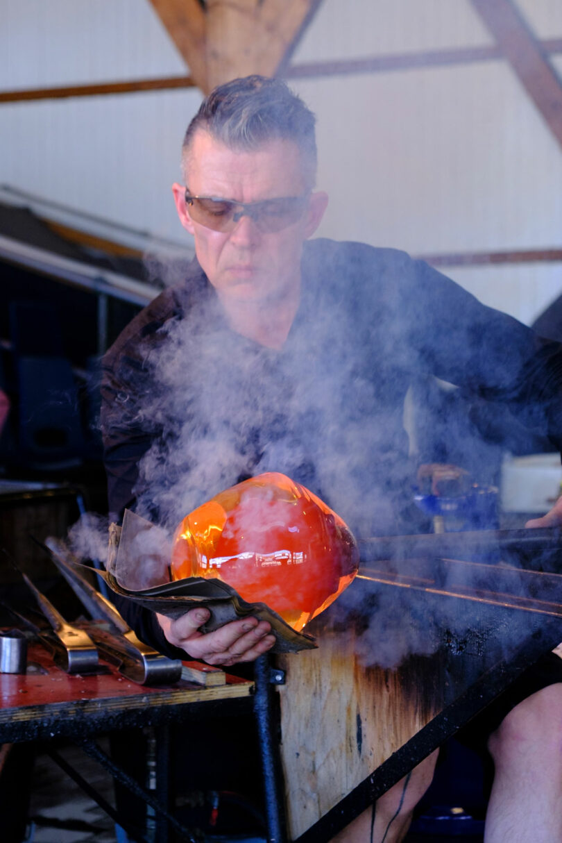 A person wearing protective glasses shapes molten glass with tools, producing steam as they work in a glassblowing studio.