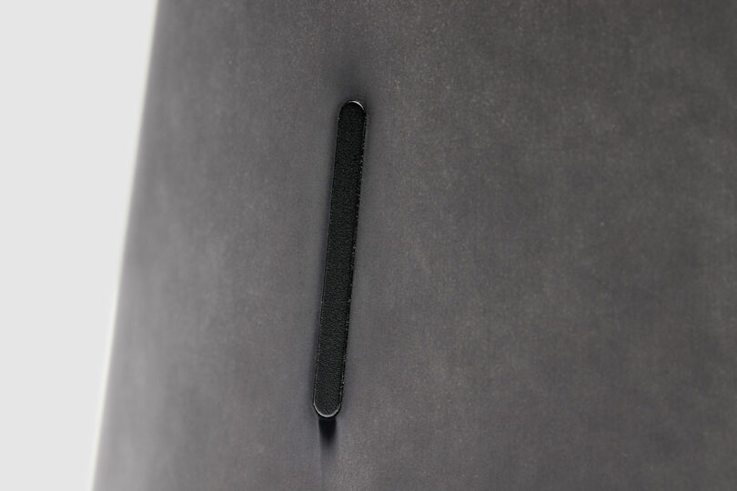 A close-up of a vertical black slit or slot on a smooth, gray surface, reminiscent of the minimalist design often seen in Bang & Olufsen products.