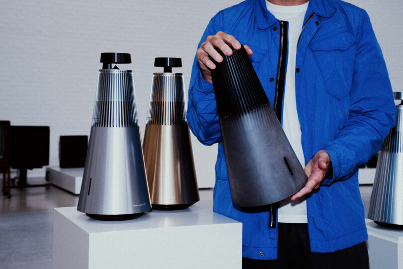 A person in a blue jacket holds a Bang & Olufsen wireless speaker; two similar cone-shaped Bang & Olufsen speakers in silver and gold are displayed on pedestals nearby.
