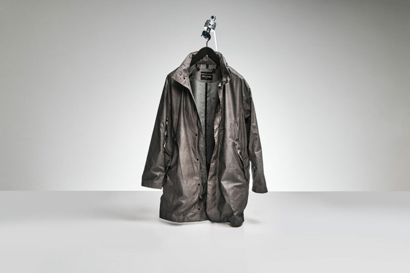 A gray, button-up raincoat hangs from a black hanger against a plain white background, with a metal clamp holding the hanger—a minimalist scene echoing the elegance of Bang & Olufsen design.