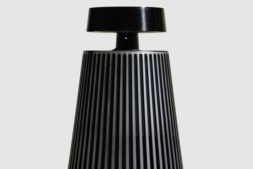 A modern, black table lamp with vertical stripes and a wide, cylindrical lampshade, inspired by Bang & Olufsen design aesthetics, set against a plain light gray background.