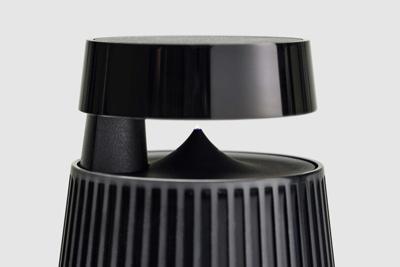 Close-up of a modern, black Bang & Olufsen wireless speaker with a cylindrical base and floating top, featuring vertical ridges on the lower section against a plain gray background.