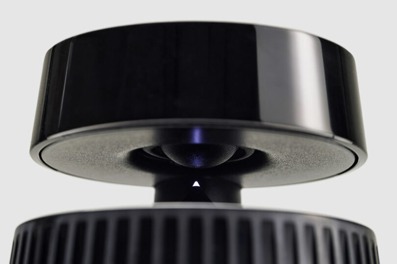 Close-up of a black Bang & Olufsen electronic device with a circular top section suspended above a base, featuring a small triangular indicator and a smooth, reflective surface.