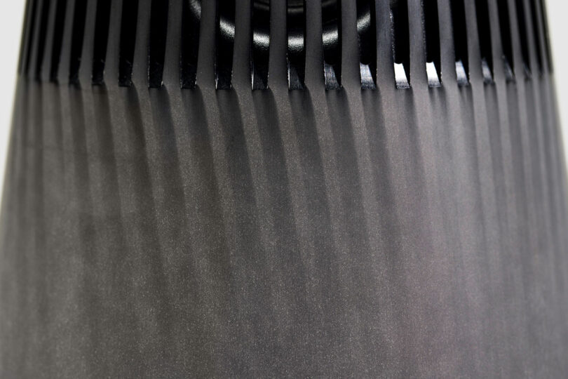 Close-up of a Bang & Olufsen black object with vertical, evenly spaced slats casting shadows on a smooth, tapered surface.