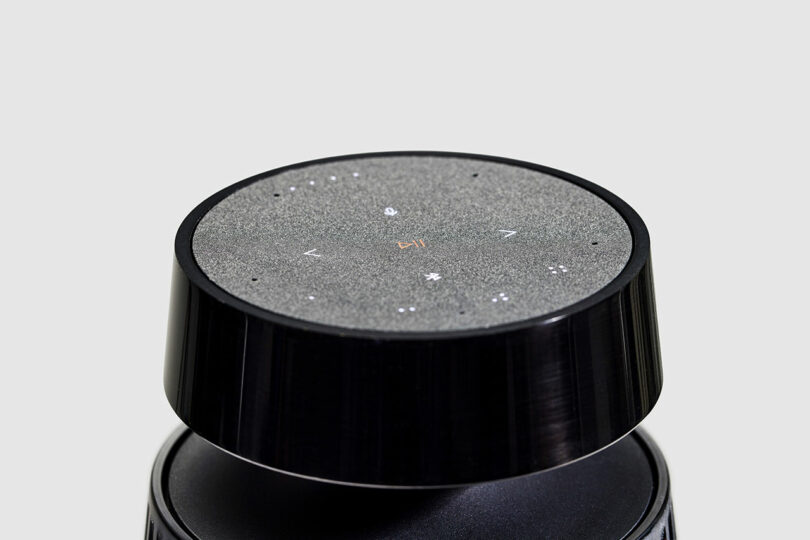 A close-up view of a black Bang & Olufsen smart speaker with a touch-sensitive surface displaying white icons and a power symbol in the center.
