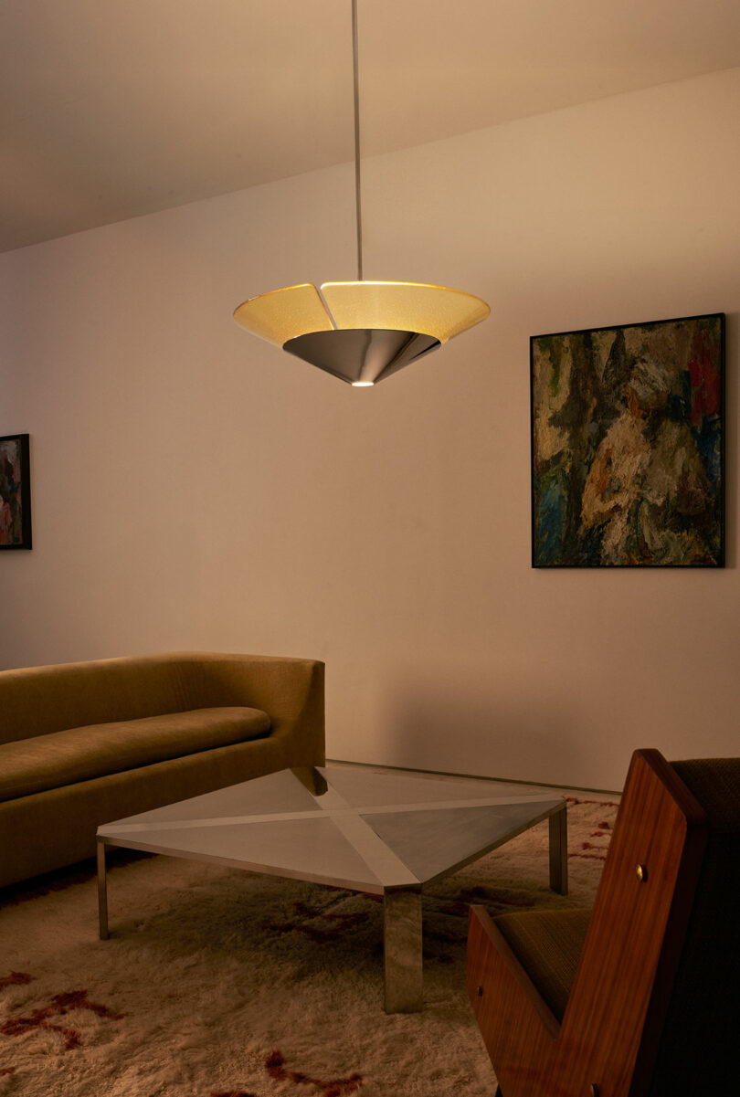 A modern living room features a triangular glass coffee table, beige curved sofa, wooden chair, abstract paintings, and a central pendant light.