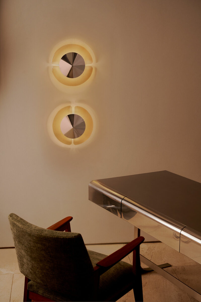 A modern office setup with a green upholstered chair, a glass and metal desk, and two circular wall lights emitting a warm glow.