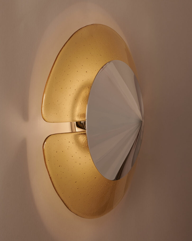 A modern wall sconce with a round, textured golden glass base and a metallic, faceted silver cover mounted on a neutral wall.