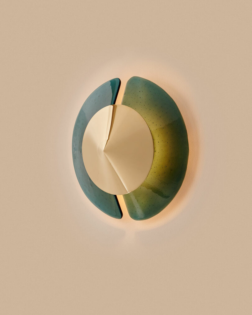 Wall light fixture with a central gold disc and two curved green glass panels on a neutral beige background.