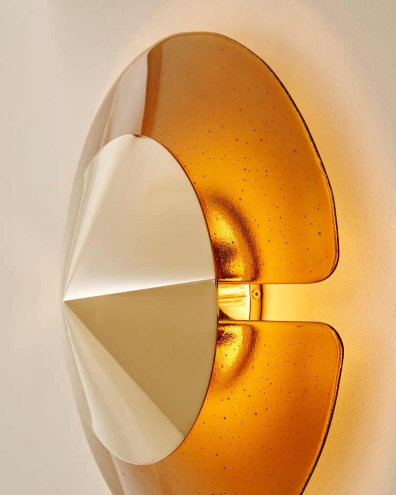 Close-up of a modern wall sconce with a metallic center and translucent amber panels, emitting a warm light against a beige wall.
