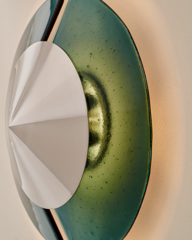 Close-up of a modern wall sconce featuring a white geometric cone intersecting a green, translucent, circular glass element mounted on a light-colored wall.