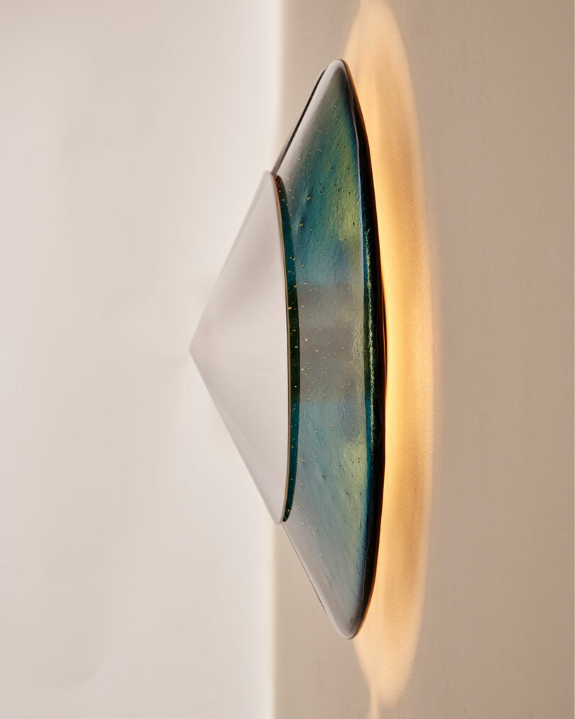 A modern wall-mounted light fixture with a conical shape, featuring frosted and translucent blue-green glass, emits a soft, warm glow onto a beige wall.
