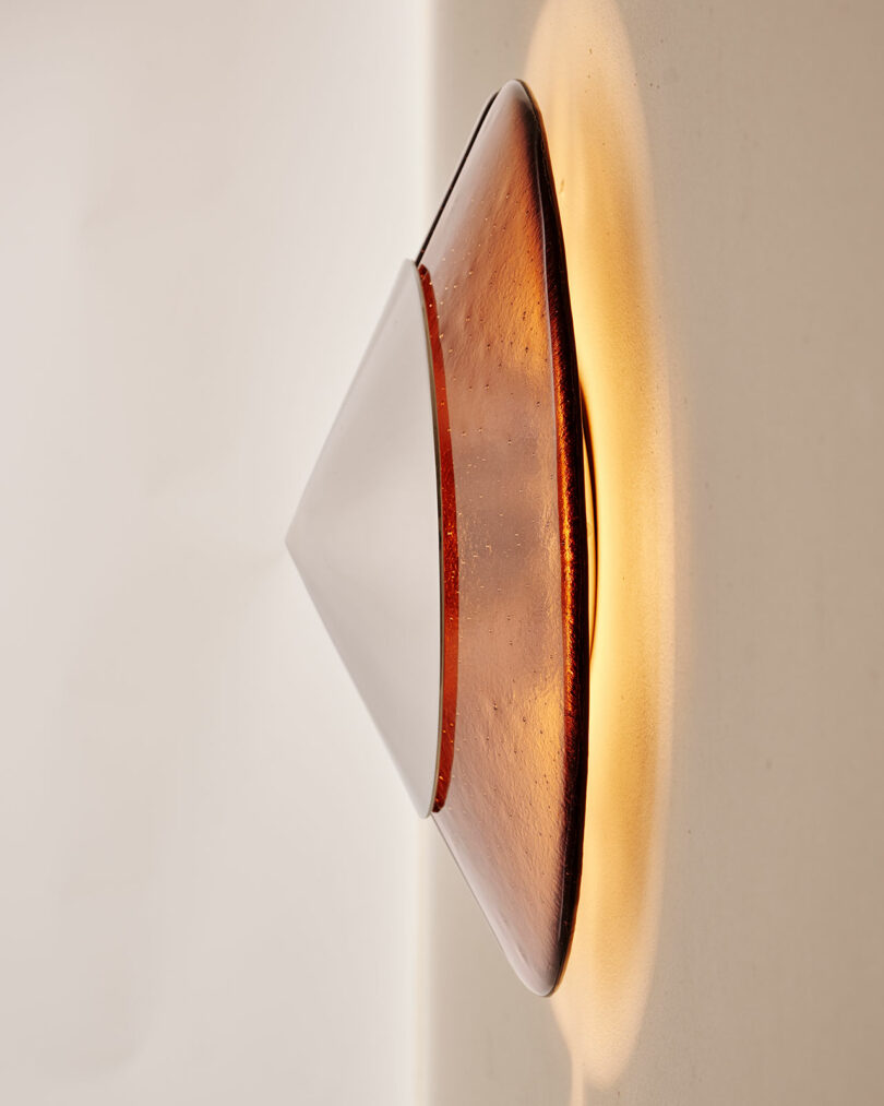 Wall-mounted light fixture with a conical, amber-colored glass shade emits a warm glow against a white wall.
