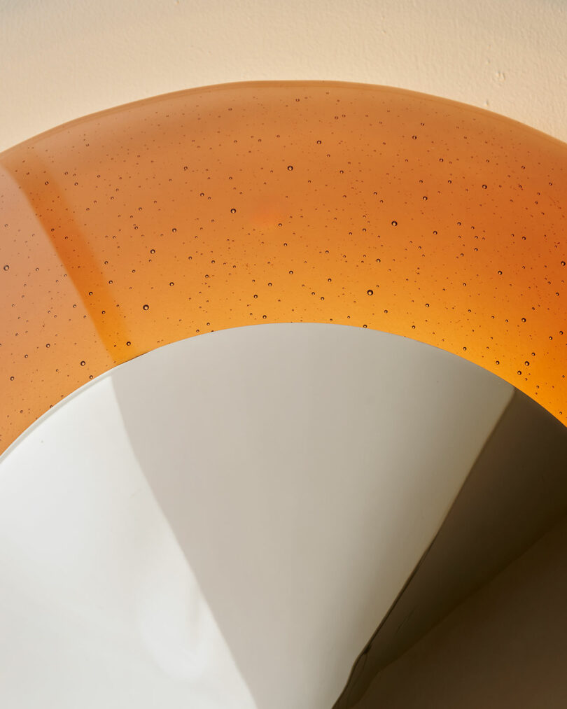 A close-up of a circular object with a glossy orange upper section featuring small bubbles and a smooth, reflective silver lower section against a beige background.