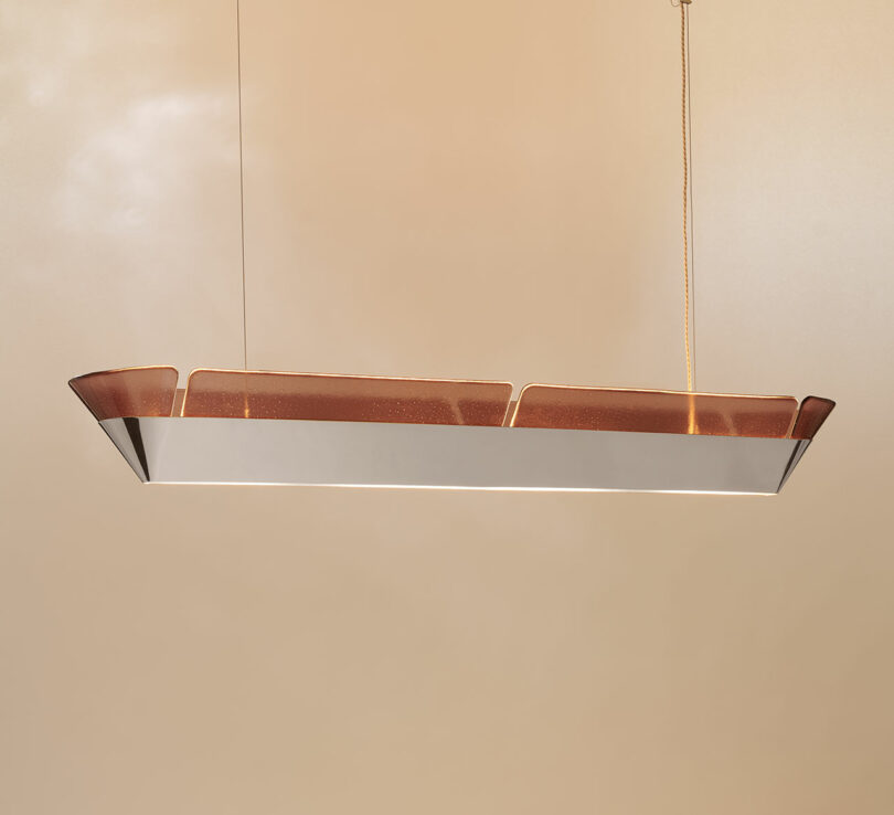 Rectangular modern pendant light with brown tinted panels and metallic base, suspended by two cables against a beige background.