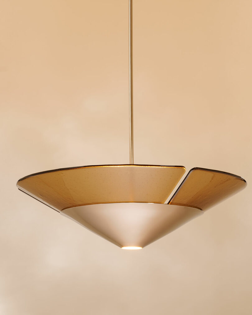 A modern pendant light with a wide, conical glass shade, suspended from a thin metal rod against a beige background.
