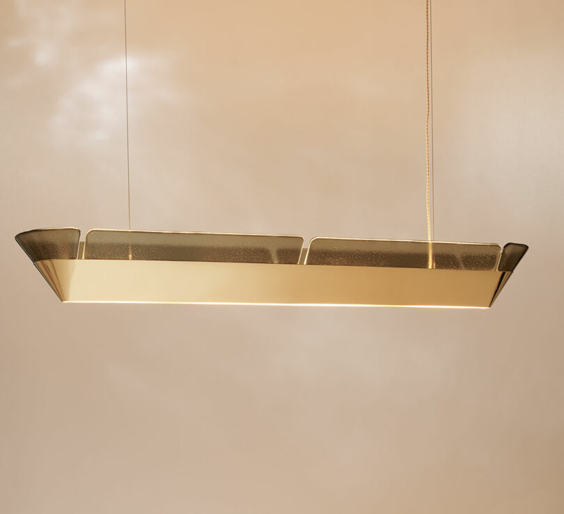 Rectangular modern pendant light with a frosted glass top and gold metallic frame, suspended by two thin cables against a beige background.