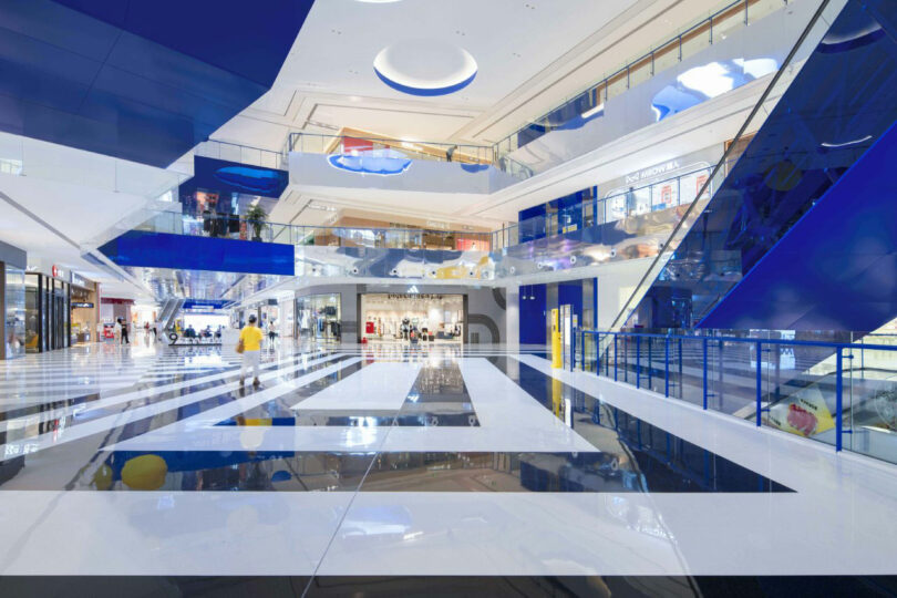 Spacious, modern shopping center Inland with reflective blue and white surfaces, glass fences and several people walking around.