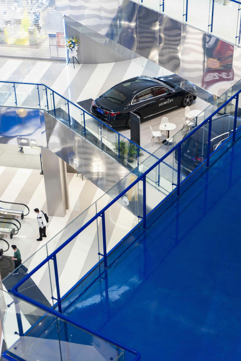 The black car is shown in a modern, multifunctional salon with blue fences, striped floors, and the person entering the final escalator.