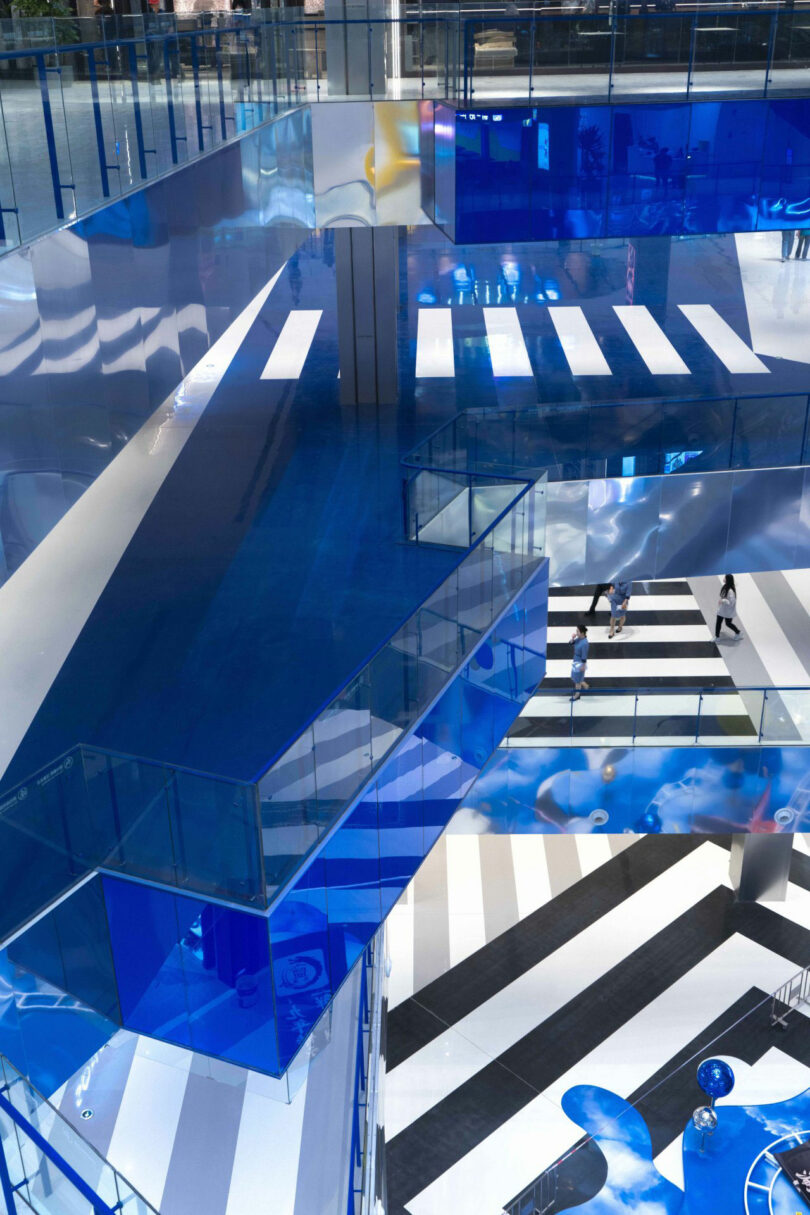 View of the inside of a modern building with bright blue and reflective surfaces, glass fences and black and white striped floors, with several people who walk below.