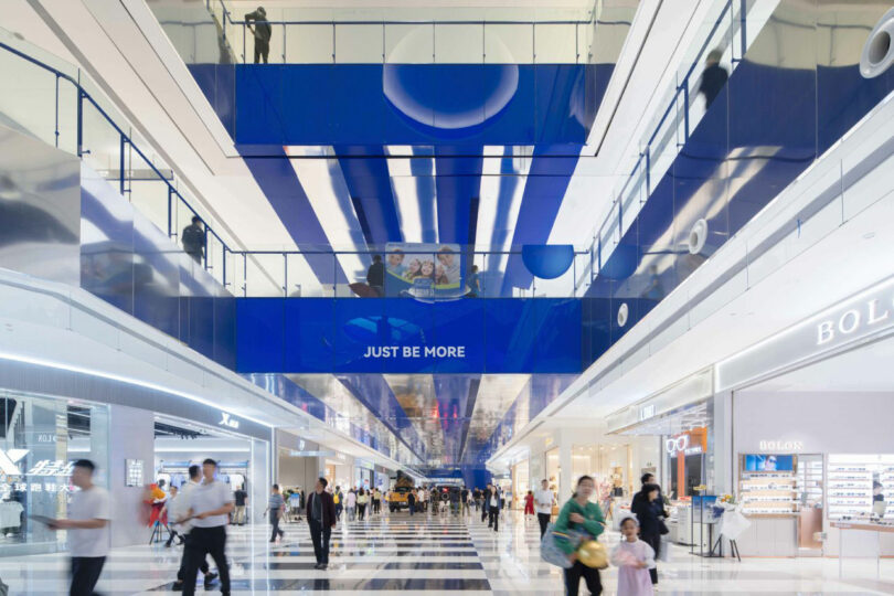 Modern shopping center Inland with reflective floors, bright lighting and banner reading in blue ceiling "Just be more." People walk and buy in a spacious corridor.