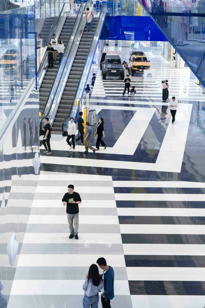 People walk and stand on a black and white striped floor in a modern shopping center; Escalators and two cars are visible in the background.