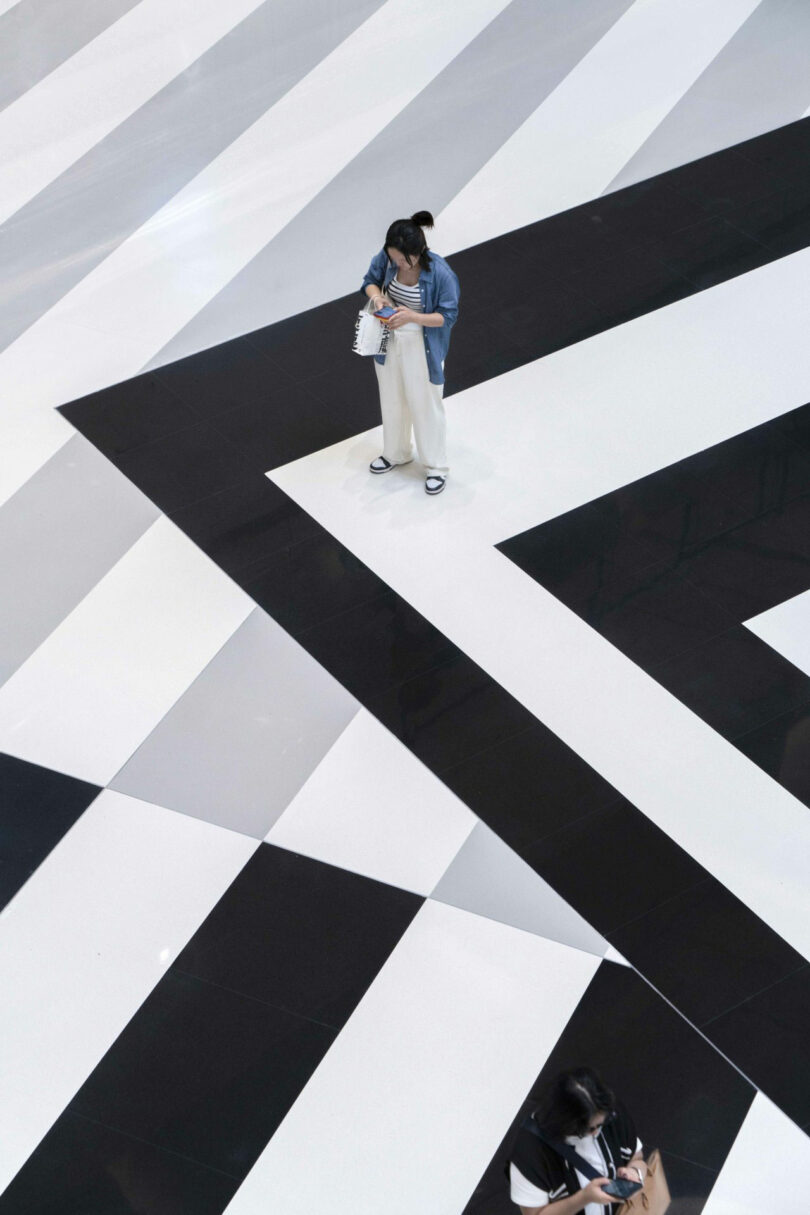 Two people stand on a large floor with bold black and white geometric patterns, observed from above.