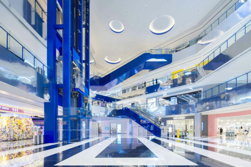 The light, modern shopping center with multiple floors, blue escalators, glass fences and ceiling lights; Stores and open spaces visible worldwide.