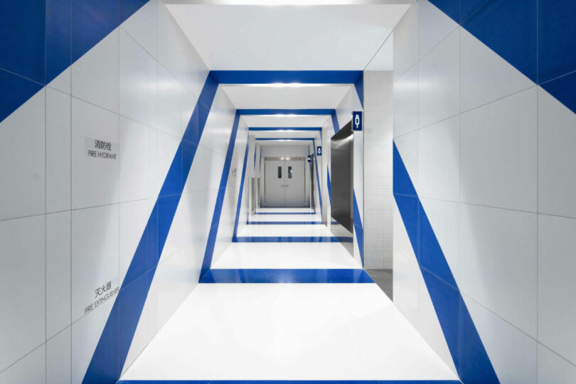Modern hallway with blue and white geometric patterns of the wall, which leads to double doors at the end; The lift is visible on the right.