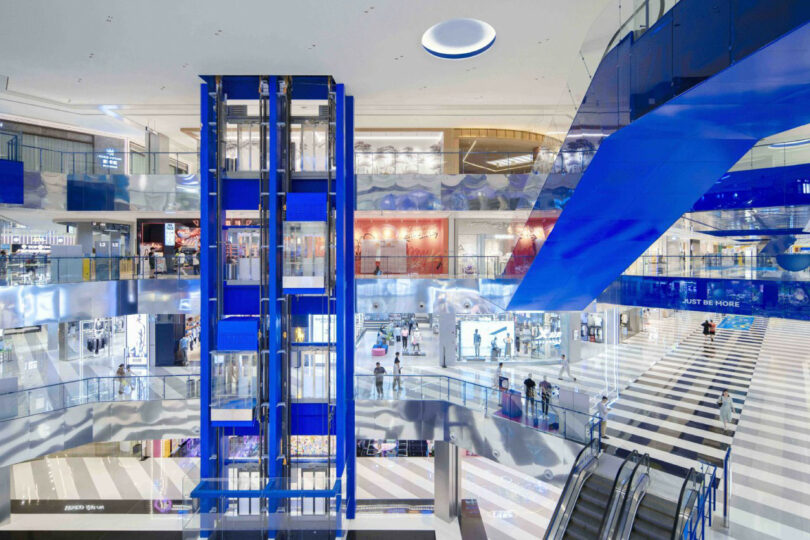 The interior of the modern shopping center with blue accents, glass elevator shafts, escalators and several people walking on striped floors.