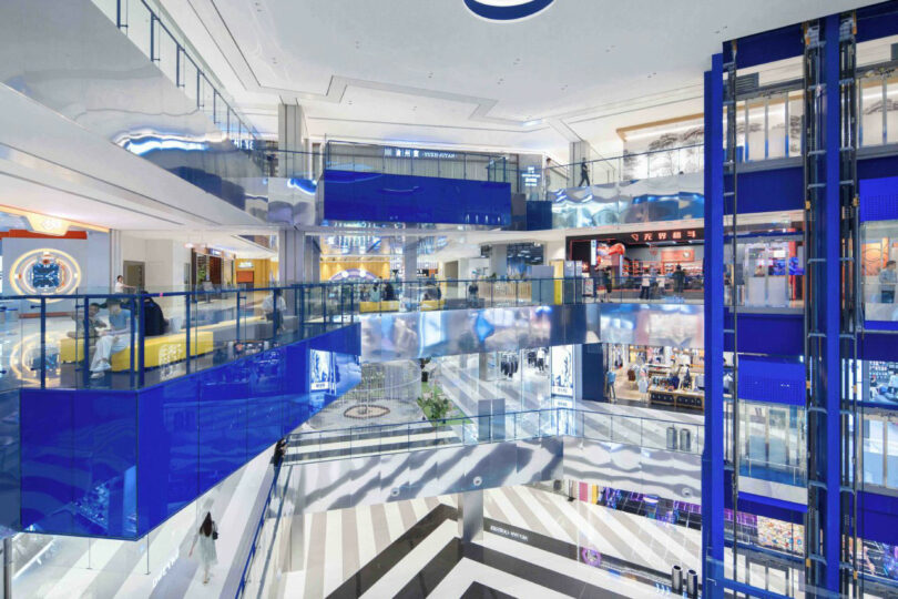 Three levels of the modern shopping center with blue and glass fences, escalators and various stores visible on each floor.