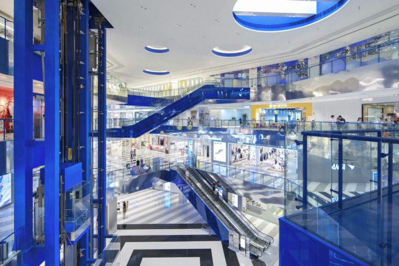 Modern shopping center Interior with multiple floors, blue glass lifts, escalators, shops and people who walk. Bright lighting and spacious design with circular ceiling lights.