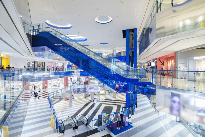 View of the interior of the modern shopping center with blue escalators, glass fences, more stores and striped floors.