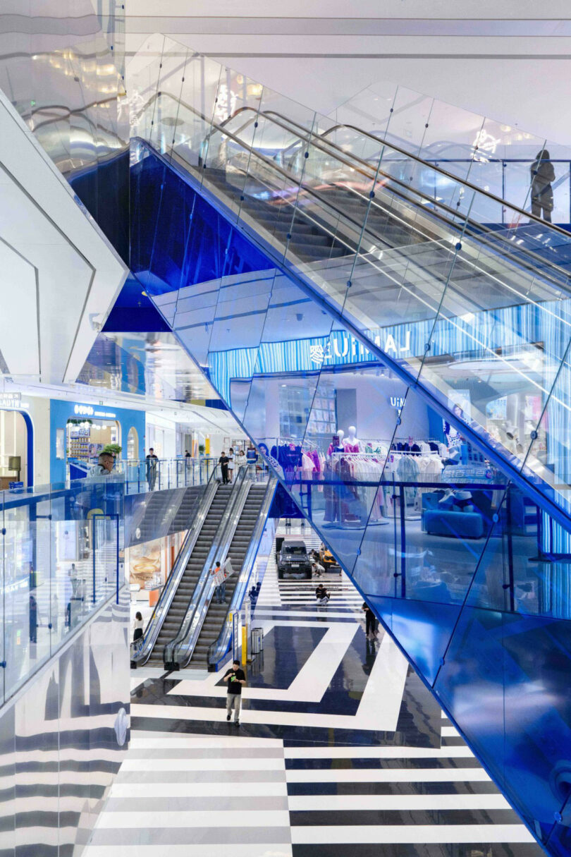 Modern shopping center Interior with blue glass accents, escalators, striped black and white floor and swimmingers visible at several levels.