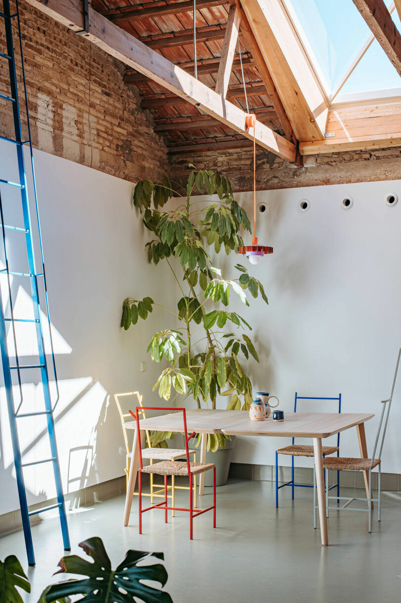 Una habitación brillante con una claraboya inclinada, vigas de ladrillo y madera expuestos, una gran planta en maceta, una mesa de madera clara y sillas coloridas.