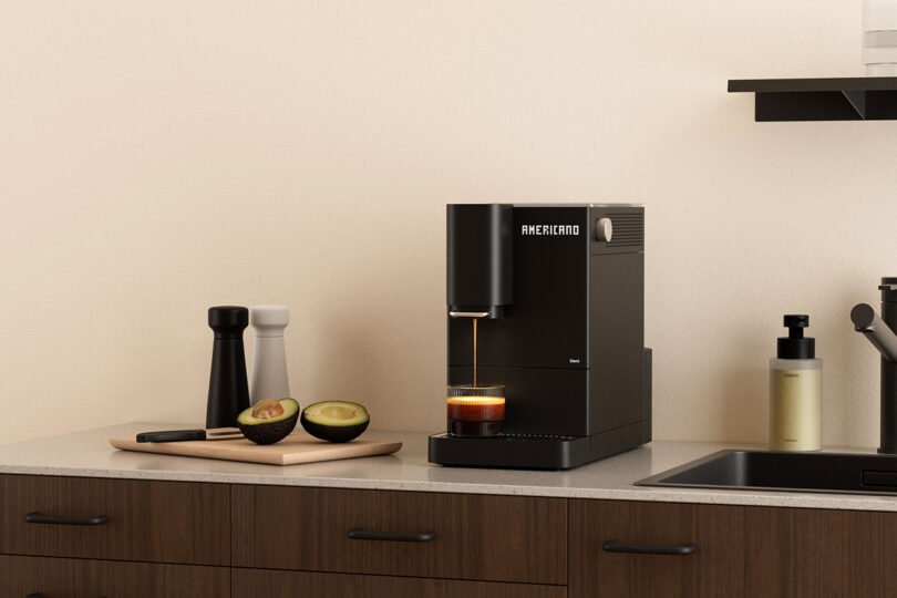 A black coffee machine labeled "AMERICANO" dispenses coffee into a cup on a kitchen counter next to a cutting board with avocado halves and salt and pepper shakers.