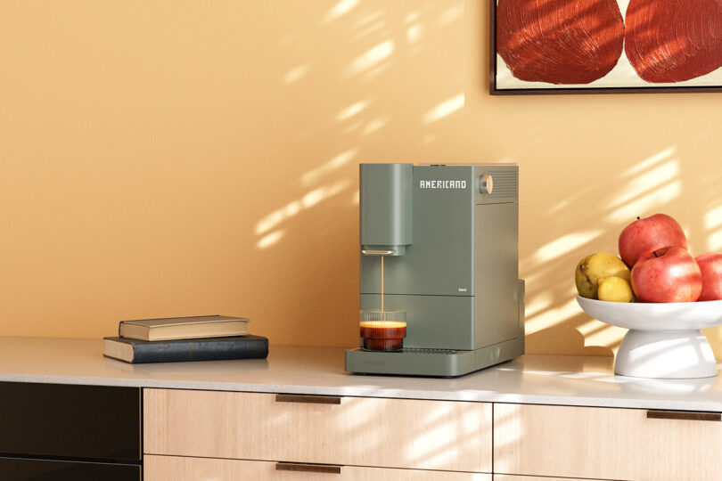 A coffee machine dispenses espresso into a glass on a kitchen counter beside a bowl of fruit and a stack of books; a painting hangs above.