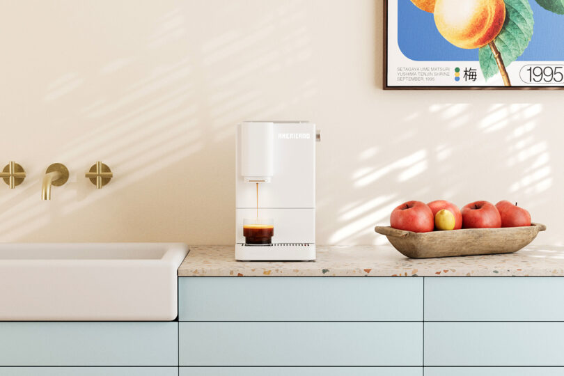 A white coffee machine brewing espresso sits on a terrazzo countertop next to a wooden bowl of apples and a sink, with a framed fruit print on the wall above.